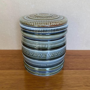 Puede incluir: Un tarro de cerámica verde y marrón con tapa. El tarro tiene un patrón decorativo de tréboles y rayas.