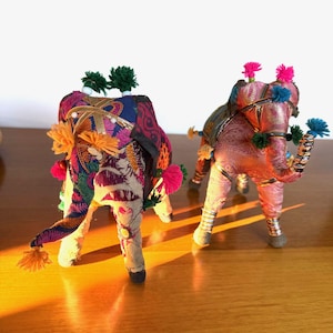May include: Two decorative elephant figurines. The elephants are made of fabric and adorned with colorful embroidery, tassels, and pom-poms. One elephant is primarily pink, the other is blue and purple. The figurines have silver accents on their legs.