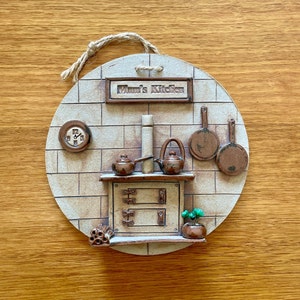 May include: A round ceramic wall plaque with a brick pattern and a stove, two teapots, two frying pans, a clock, and a plant. The plaque has the text "Mum's Kitchen" on it.