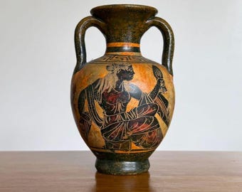 May include: A vintage, two-handled amphora vase with a painted scene of a seated figure holding a mirror. The vase features a dark green base and handles, with orange and black detailing. The figure is depicted in black and red.