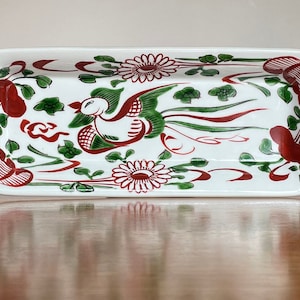 Op de afbeelding: Een rechthoekige serveerschaal van wit keramiek met een rood en groen bloemmotief. Het ontwerp toont een gestileerde vogel met een lange staart en bloemen.