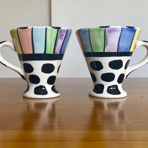 May include: Two white ceramic mugs with black polka dots and colorful stripes. The mugs have a flared shape and a handle.