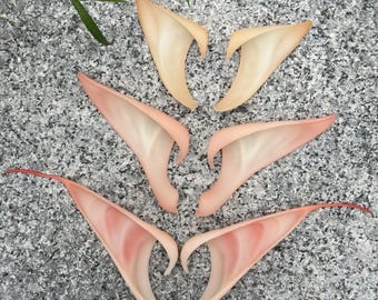 May include: Three pairs of pointed ear prosthetics in varying skin tones, ranging from a light beige to a soft pink and a deeper reddish-pink hue. Each pair features a curved, elongated shape designed to fit over the human ear structure.