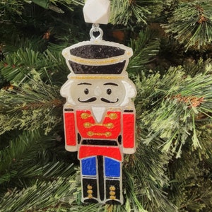 May include: A red and blue nutcracker ornament with gold accents and glitter. The ornament is hanging from a white and gold beaded string.