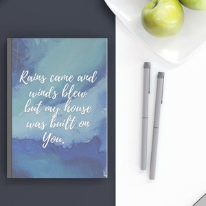 Puede incluir: Un cuaderno de acuarela azul y blanco con la cita "Las lluvias llegaron y los vientos soplaron, pero mi casa fue construida sobre ti." El cuaderno está sobre un escritorio con un portátil, gafas, bolígrafos y manzanas.