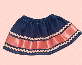 May include: A navy blue skirt with a wide coral satin band and a wavy white trim. The skirt has an elastic waistband and is likely for a child. The skirt is set against a light pink background with white polka dots.