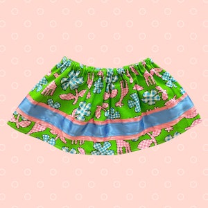 May include: A green skirt with an elastic waistband, featuring a pattern of pink giraffes, blue and white checkered flowers, and pink leaves. The skirt has a blue ribbon and a pink ribbon trim.