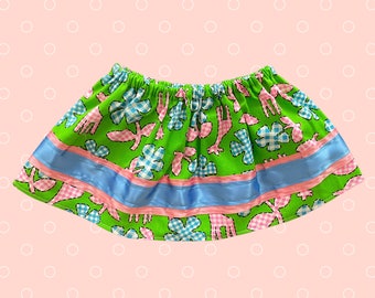 May include: A green skirt with an elastic waistband, featuring a pattern of pink giraffes, blue and white checkered flowers, and pink leaves. The skirt has a blue ribbon and a pink ribbon trim.
