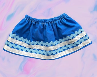 May include: A blue skirt with an elastic waistband. The skirt features white and light blue ribbon trim with a wavy design. The skirt is made of a solid blue fabric.