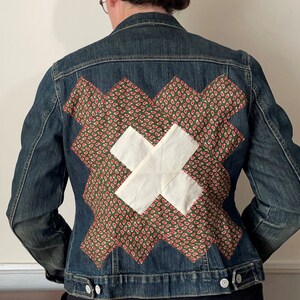 May include: A dark wash denim jacket with a patchwork design on the back. The patchwork features a green paisley pattern with red accents and a white cross design. The jacket has silver buttons and a classic collar.