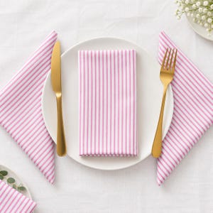May include: A white ceramic dinner plate centred with a folded pink and white striped cloth napkin. A gold-toned metal dinner knife is placed on the left side of the plate, and a gold-toned metal fork is placed on the right side of the plate.