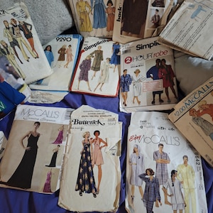 May include: A collection of vintage sewing pattern envelopes from brands including Butterick, Simplicity, and McCall's. The paper envelopes feature various illustrations of clothing designs, including dresses, robes, and loungewear, with printed text.