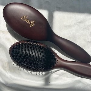 Peut inclure: Deux brosses à cheveux ovales avec des manches et des dos en bois brun foncé. La brosse supérieure porte le prénom Emily gravé en lettres dorées sur le dos. La brosse inférieure présente une densité de poils noirs sur une base brune.