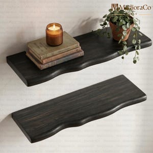 May include: Two dark stained wooden floating shelves with a live edge design. One shelf holds a stack of vintage books, a lit candle in a glass jar, and a potted plant. The shelves are mounted on a light-colored wall.