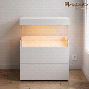 May include: A white wooden table with a built-in display case, illuminated from within. The table has a drawer at the bottom and a top section with a light. The table is made of wood and has a modern design.
