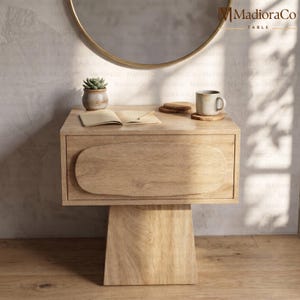 Puede incluir: Una mesita de noche de madera clara con un cajón y una base de pedestal. La mesita de noche tiene un diseño redondeado en el cajón. Una pequeña suculenta en una maceta, un libro y una taza están encima. Un espejo dorado está en el fondo.
