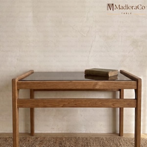 May include: A wooden table with a dark glass top and a lower shelf. The table has a rectangular shape and a natural wood finish. A small, aged book rests on the tabletop. The table is branded with the text "MADIORA CO TABLE".