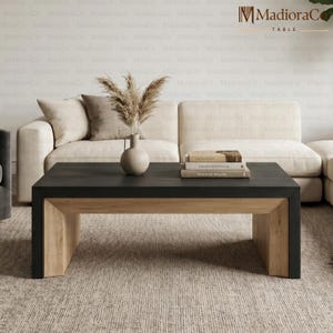 May include: A rectangular coffee table featuring a matte black top surface and light natural wood grain legs. A beige ceramic vase containing dried pampas grass and a stack of three books are placed on the center of the table surface.