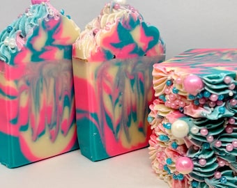 May include: Three bars of handcrafted soap featuring a vibrant swirl of hot pink, bright turquoise, and cream colors. The tops are decorated with piped soap frosting, small white and pink spherical beads, and larger pearl-like accents.