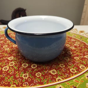 May include: A blue enamel chamber pot with a white interior and a black rim. The pot has a single handle and is sitting on a red and green floral tablecloth.