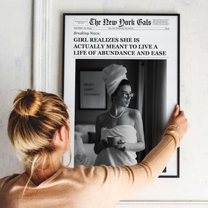 Pu&ograve; includere: Un titolo di giornale in bianco e nero recita "Breaking News: Girl Realizes She Is Actually Meant To Live A Life Of Abundance And Ease". Una donna con un asciugamano bianco e occhiali da sole tiene una tazza di caff&egrave;. Il testo "Enjoy Every Moment" &egrave; visibile su una stampa incorniciata dietro di lei.