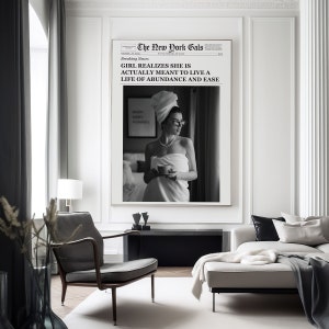 Pu&ograve; includere: Foto in bianco e nero di una donna avvolta in un asciugamano, che tiene una tazza di caff&egrave;. La foto &egrave; incorniciata e appesa a una parete in un soggiorno moderno. Il testo sulla foto recita "The New York Gals" e "Breaking News: GIRL REALIZES SHE IS ACTUALLY MEANT TO LIVE A LIFE OF ABUNDANCE AND EASE."