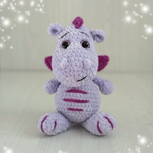 Puede incluir: Un juguete de peluche de drag&oacute;n de crochet morado con detalles en rosa y ojos negros. El drag&oacute;n est&aacute; sentado sobre una superficie blanca.