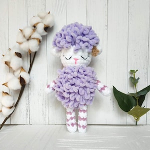 May include: A crocheted purple and white sheep plush toy with a sweet expression. The toy has a fluffy texture and is standing on a white surface.