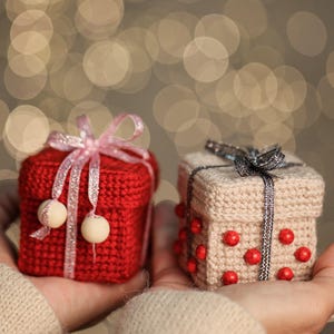 May include: Two small, crocheted gift boxes are held in open hands. One is red with a pink ribbon and white bead accents. The other is beige with a silver ribbon and red bead accents. The background is a soft bokeh of warm lights.