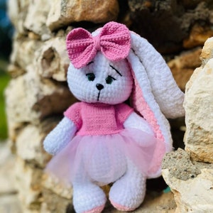 Op de afbeelding: Een witte gehaakte bunny met roze oren, een roze strik en een roze tutu. De bunny draagt een roze shirt en zit op een rots.