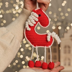 May include: A handmade red crochet horse ornament with white mane, tail, and trim. The horse has a small blue star on its head and red pom-poms for feet. The ornament is held up by a white string.
