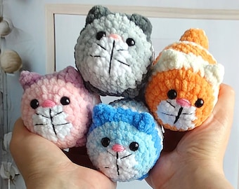 Könnte beinhalten: Vier gehäkelte Katzenspielzeuge in verschiedenen Farben: grau, rosa, orange und blau. Jede Katze hat ein weißes Gesicht mit schwarzen Augen und einer schwarzen Nase.