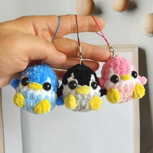 May include: Three small, crocheted penguin keychains in blue, black and white, and pink. Each penguin has black button eyes and yellow feet. The keychains are held by a hand against a neutral background.