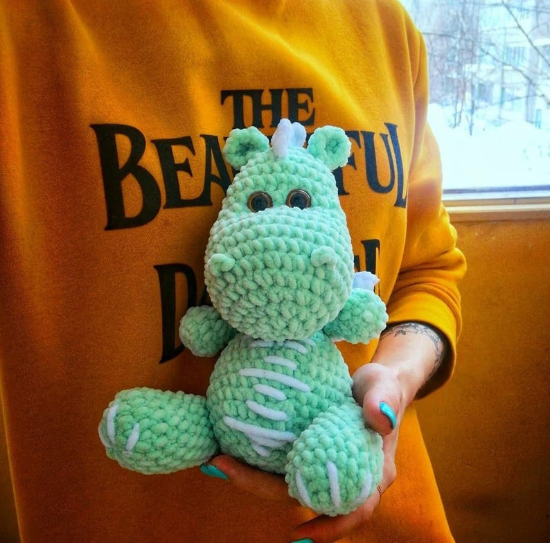 Puede incluir: Un juguete de drag&oacute;n de crochet de color verde claro con detalles blancos. El drag&oacute;n tiene un peque&ntilde;o cuerno en la cabeza y ojos grandes y redondos. El juguete est&aacute; siendo sostenido por una persona que lleva una sudadera amarilla con el texto "The Beautiful Day" impreso en ella.
