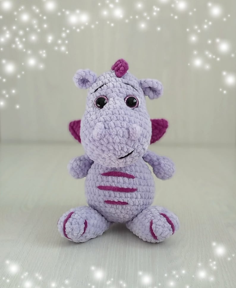 Puede incluir: Un juguete de peluche de drag&oacute;n de crochet morado con detalles en rosa y ojos negros. El drag&oacute;n est&aacute; sentado sobre una superficie blanca.