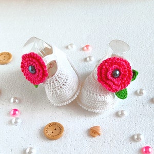 Pu&ograve; includere: Due scarpine da beb&egrave; in crochet bianco con decorazioni di fiori rosa. Ogni fiore ha un bottone nero al centro.
