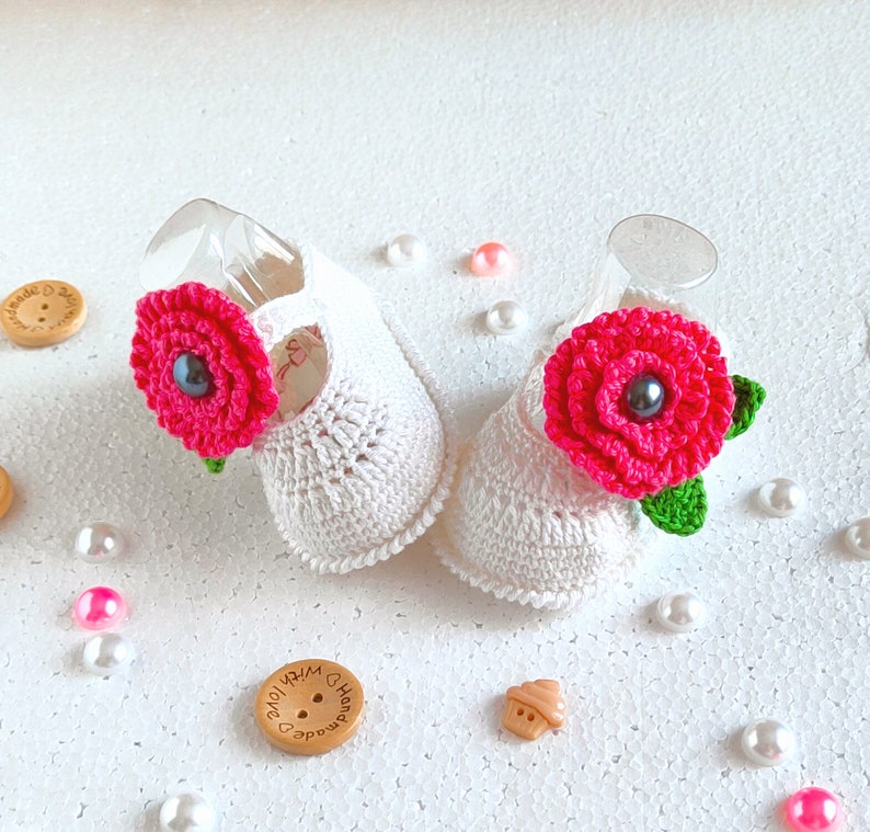 Pu&ograve; includere: Due scarpine da beb&egrave; in crochet bianco con decorazioni di fiori rosa. Ogni fiore ha un bottone nero al centro.
