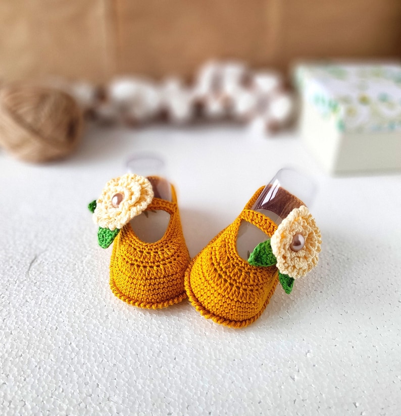 Pu&ograve; includere: Un paio di scarpine per neonati in crochet giallo con un fiocco di fiori bianco su ogni scarpa.