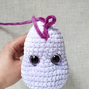 Puede incluir: Un juguete de peluche tejido a crochet en forma de mancha p&uacute;rpura con detalles de hilo rosa y ojos negros con bordes rosas.