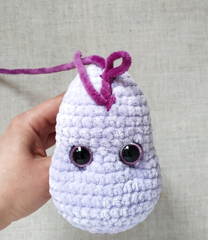 Puede incluir: Un juguete de peluche tejido a crochet en forma de mancha p&uacute;rpura con detalles de hilo rosa y ojos negros con bordes rosas.