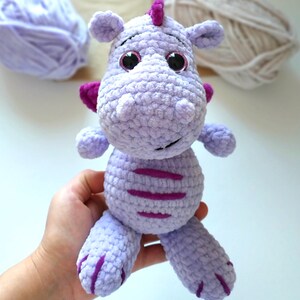 Puede incluir: Un juguete de peluche de drag&oacute;n de crochet morado con ojos rosas y un cuerno morado. El drag&oacute;n tiene un patr&oacute;n de rayas en su cuerpo.