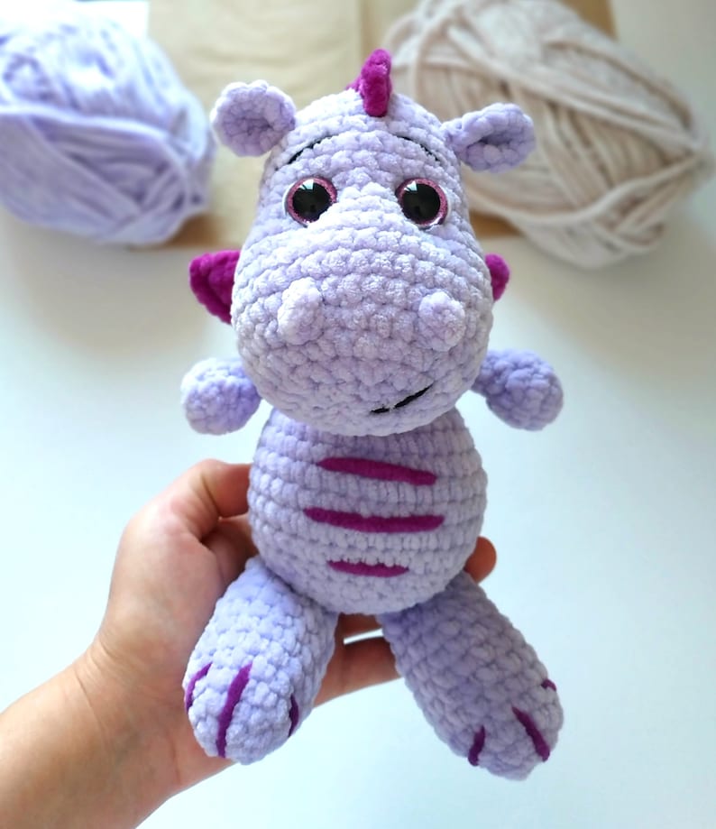 Puede incluir: Un juguete de peluche de drag&oacute;n de crochet morado con ojos rosas y un cuerno morado. El drag&oacute;n tiene un patr&oacute;n de rayas en su cuerpo.