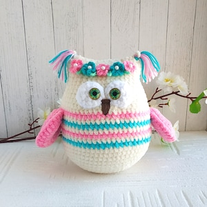 May include: A crocheted owl plush toy with pink and blue stripes, a white face, green eyes, and a flower crown with pink and blue flowers.