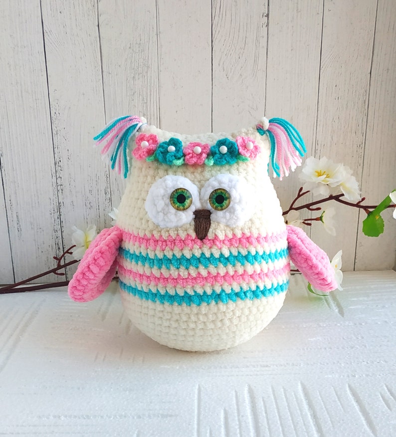 May include: A crocheted owl plush toy with pink and blue stripes, a white face, green eyes, and a flower crown with pink and blue flowers.