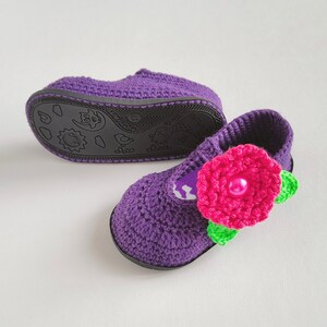 Pu&ograve; includere: Scarpe per neonati in crochet viola con un fiore rosa e foglie verdi. Le scarpe hanno una suola nera con un design a cartoni animati.