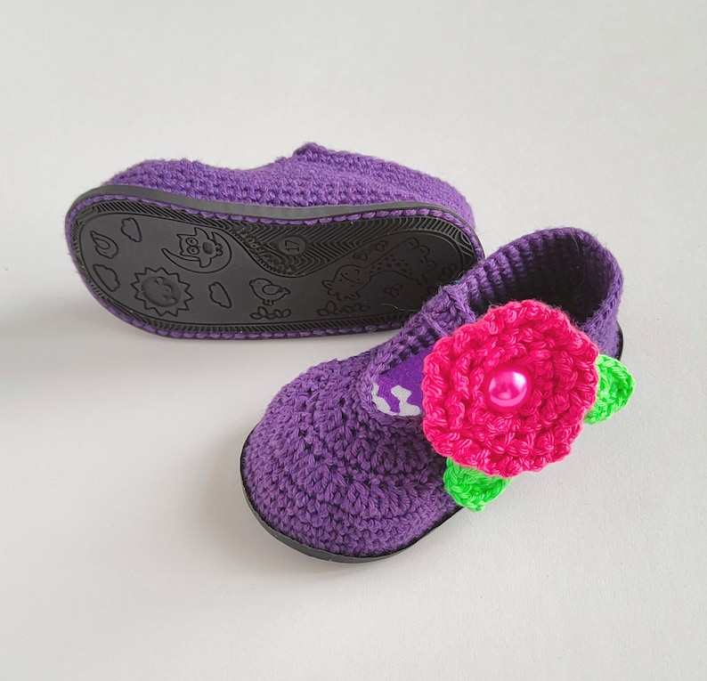 Pu&ograve; includere: Scarpe per neonati in crochet viola con un fiore rosa e foglie verdi. Le scarpe hanno una suola nera con un design a cartoni animati.
