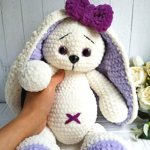 Op de afbeelding: Een witte gehaakte bunny met paarse oren en een paarse strik. De bunny heeft zwarte ogen en een paarse geborduurde X op zijn borst.