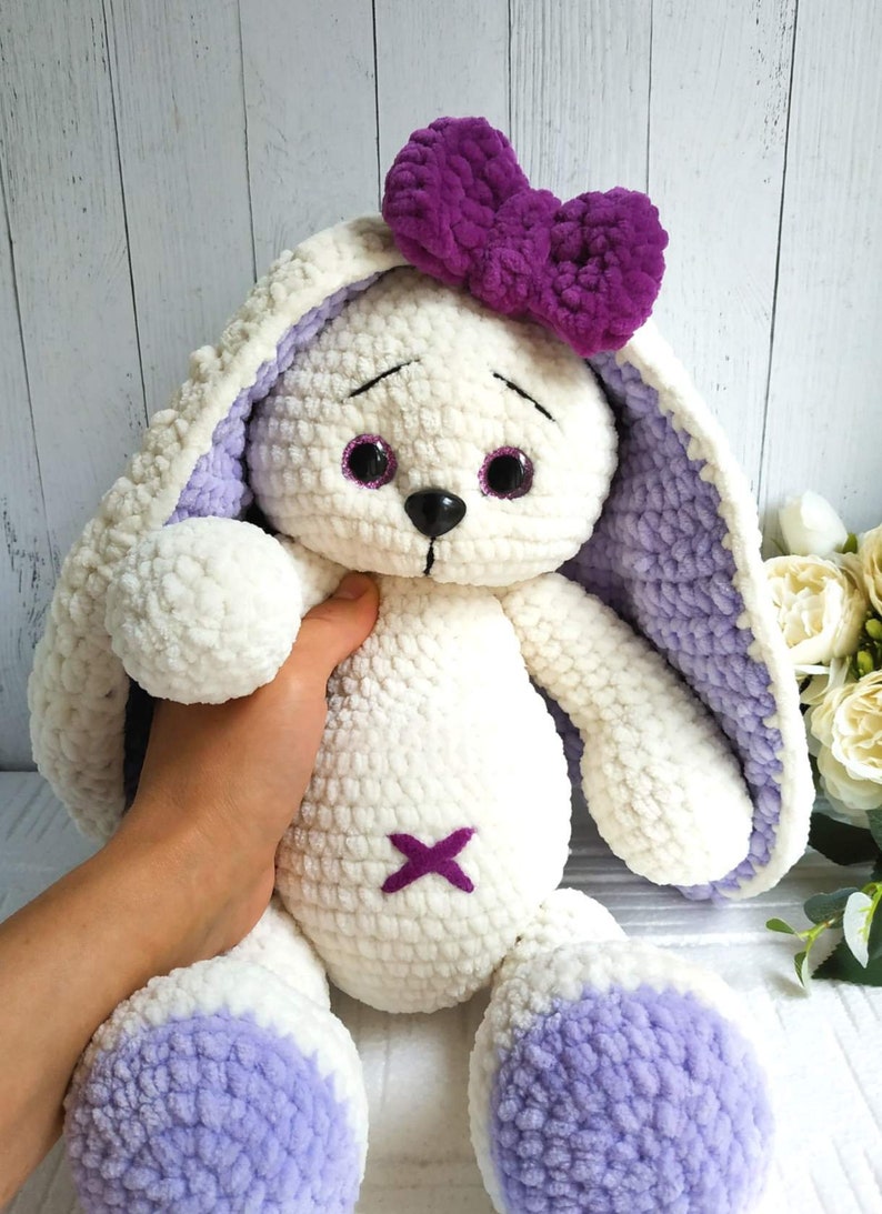 Op de afbeelding: Een witte gehaakte bunny met paarse oren en een paarse strik. De bunny heeft zwarte ogen en een paarse geborduurde X op zijn borst.