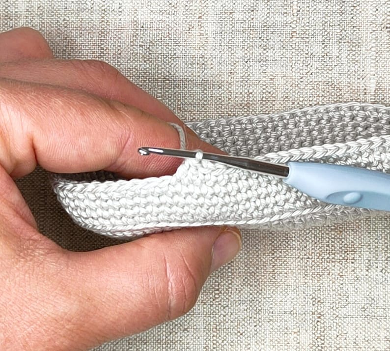 Pu&ograve; includere: Primo piano di una persona che lavora a uncinetto con un filo di colore grigio chiaro e un uncinetto blu. L'uncinetto &egrave; inserito in un punto sul bordo di un pezzo lavorato a uncinetto.