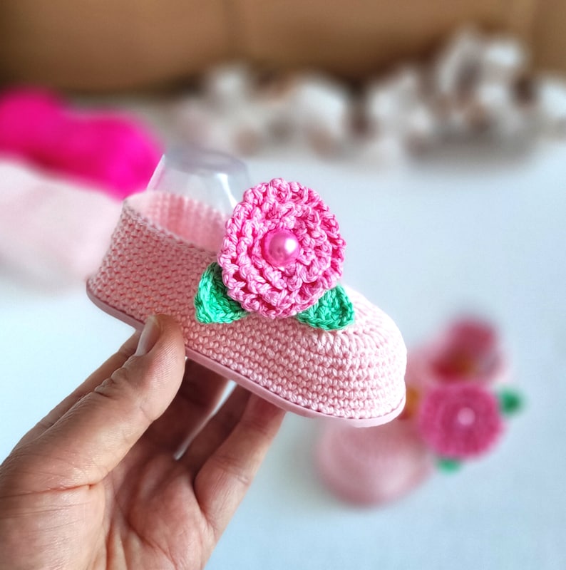 Pu&ograve; includere: Scarpetta per neonato in crochet rosa con un fiore rosa e foglie verdi. Il fiore ha un centro perlato.